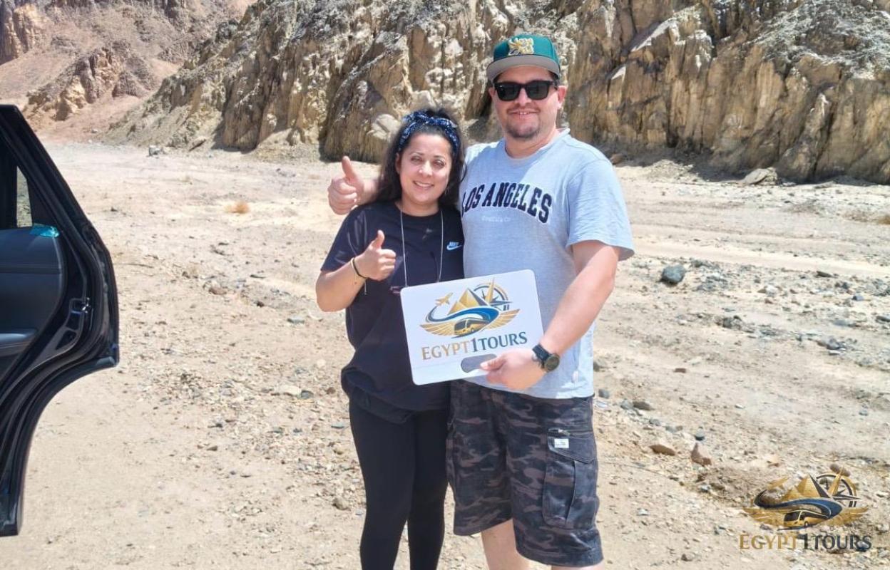 "Happy international clients on the Eastern Desert road from Luxor to Hurghada holding the Egypt1Tours logo sign, proving our private transfer service reliability."
